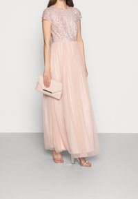 Largo vestido color rosa empolvado con un corpiño bordado de lentejuelas, mangas cortas y una falda de tul fluida. Combinado con un bolso de mano rosa claro y tacones transparentes.