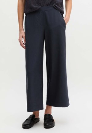 Kobieta ubrana w czarne szerokie spodnie do kostek z bocznymi kieszeniami oraz czarne wsuwane mokasyny na jednolitym białym tle.