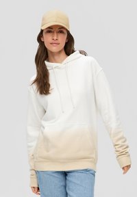 Sudadera con degradado beige claro, con un bolsillo canguro y cordones. Combinada con una gorra beige. Presenta una textura de tela suave.