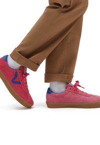 Red suede sneakers with a blue heel, featuring a round toe, flat rubber sole, and matching laces. Pants are brown and slightly rolled.