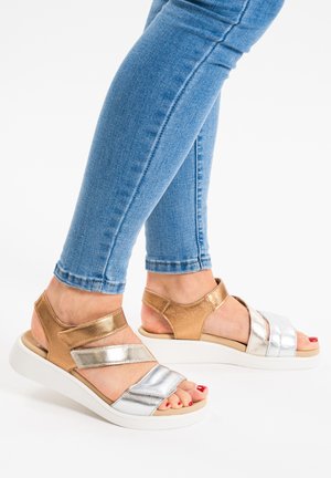 Sandaletter med remme i metallic guld og sølv, med en hvid sål og justerbare stropper, parret med lyseblå jeans.