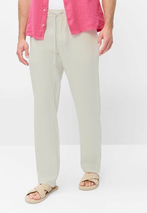 Man draagt lichtbeige broek met trekkoord, beige sandalen met kruisbandjes, een wit overhemd en een open roze overhemd met knopen.