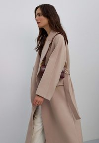 Lange, beige jas met een losse pasvorm, grote kraag en gestikte details; gecombineerd met een bordeauxrode schoudertas. Gladde stof, minimalistisch ontwerp.