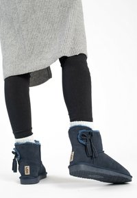 Navy blue ankle boots made of suede, featuring a tassel accent, a textured rubber sole, and a soft, light-colored lining.
