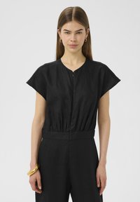 Combinaison noire en lin avec décolleté en V et manches courtes, dotée d'une taille cintrée. Le modèle porte un bracelet en or à son poignet. Design minimaliste.