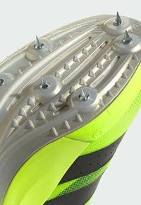 Semelle de chaussure de trail avec six pointes métalliques et plusieurs petits picots en caoutchouc, dessus néon jaune et noir visible sur le côté.