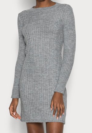 Femme portant une robe moulante en maille côtelée grise avec des détails en maille torsadée et des manches longues, debout devant un fond neutre.