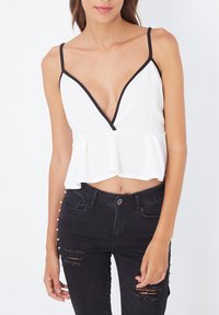 Top peplum blanco con escote en V profundo, acentuado con borde negro. Combinado con jeans ajustados negros con detalles rasgados y adornos de perlas.