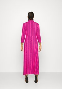 Fuchsia lang kjole med chevronmønster, 3/4-ærmer og en figursyet talje. Sorte hælede sandaler fuldender outfittet.