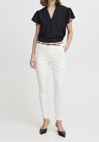 Schwarze kurzärmelige Bluse mit V-Ausschnitt und strukturiertem Muster, kombiniert mit weißen schmal geschnittenen Hosen und einem braunen Gürtel. Schwarze spitze Pumps.