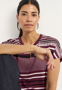 Mørkerød- og hvidstribet t-shirt med korte ærmer, båret med mørk denim. Et guld kædearmbånd pryder håndleddet. Glat stofstruktur.