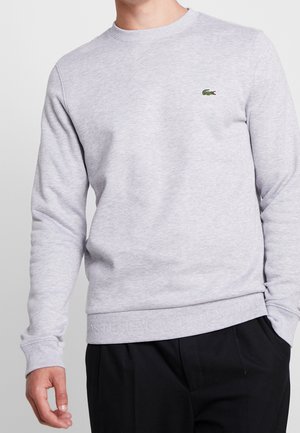 Mand iført en lysegrå langærmet sweatshirt med et lille grønt krokodillelogo på brystet og sorte bukser.