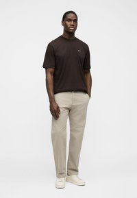 Homme portant un t-shirt à manches courtes marron foncé, un pantalon beige et des baskets blanches, debout devant un fond clair uni.