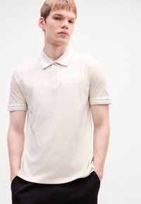 Cămașă polo de culoare crem, dintr-un material neted, cu guler și manșete ribbed albe, plachetă cu trei nasturi și detalii subtile cu siglă.