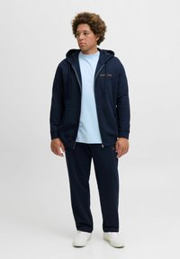 Marine blauwe zip-up hoodie met trekkoorden, gecombineerd met een lichtblauw T-shirt en marineblauwe joggingbroek. Witte sneakers maken de outfit compleet.