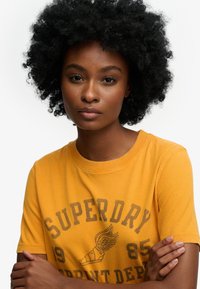 Geel katoenen T-shirt met korte mouwen. Heeft een bedrukte grafische afbeelding in bruin, inclusief een ontwerp van een gevleugelde sandaal en de tekst "SUPERDRY 1985".