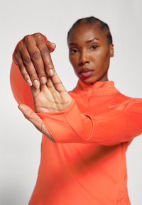 Nike langarmtop in feloranje, met een hoge kraag en aansluitende mouwen, voorzien van een gladde textuur en duimgaten voor extra comfort.