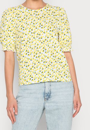 Frau trägt eine weiße Bluse mit gelbem Zitronenprint und aufgepufften kurzen Ärmeln, kombiniert mit hellblauen Jeans.