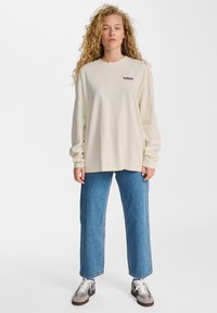 Langärmliges cremefarbenes T-Shirt mit kleinem Logo, kombiniert mit lockeren blauen Jeans und grauen Sneakers mit weißen Akzenten.