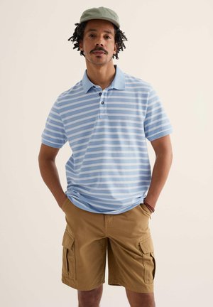 Mann trägt hellblau-weiß gestreiftes Poloshirt, khakifarbene Cargo-Shorts, grüne Kappe und braunes Armband, steht mit den Händen in den Taschen.