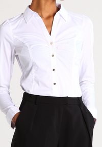 Weißes Langarm-Hemd mit Knopfverschluss und Kragen, ausgestattet mit schwarzen Knöpfen und feinen Rüschendetails an den Schultern, kombiniert mit schwarzen Hosen.