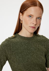 Grøn strikket sweater med en tekstureret, glitrende finish og guldknap-detaljer på skuldrene. Har en rund halsudskæring og korte ærmer.