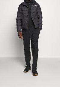 Man wearing black puffer jacket, black zip-up sweater, black pants, and black sneakers with brown soles against a plain light background.