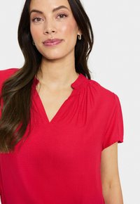 Blusa rossa con scollo a V e maniche corte leggermente arricciate. Presenta una sottile texture e arricciature nel punto collo.