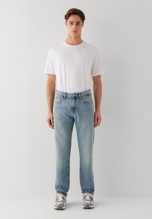 Ein junger Mann steht frontal mit einem schlichten weißen T-Shirt, hellblauen Jeans und grauen sowie weißen Turnschuhen vor einem weißen Hintergrund.