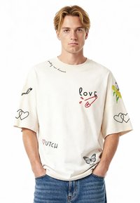 Kreminė medvilninė marškinė su per dideliu siluetu. Yra įvairių grafikų atspaudų, įskaitant širdeles, žodį "meilė" ir drugelį ant rankovių.