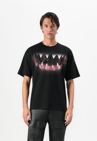 Man wearing black T-shirt with large graphic of sharp teeth and gums, paired with black leather pants, standing against white background.