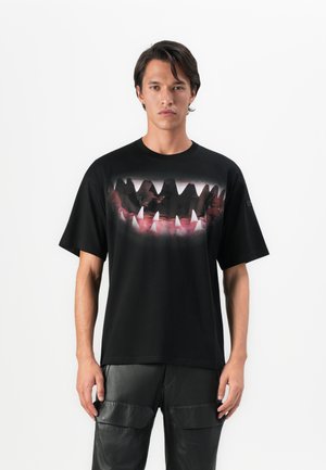 Mann in schwarzem T-Shirt mit großem Grafikdruck von scharfen Zähnen und Zahnfleisch, kombiniert mit schwarzer Lederhose, steht vor weißem Hintergrund.