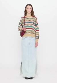 Maglione a maglia con motivi geometrici colorati, abbinato a una gonna maxi in denim chiaro con spacchi laterali. Gli accessori includono una borsa bordeaux.