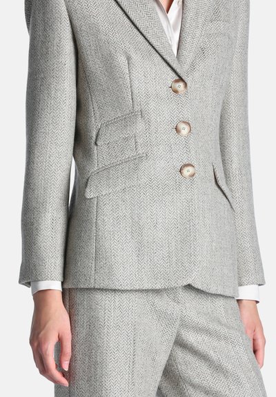 Femme portant un costume en chevrons gris clair avec trois boutons, des poches à rabat et un pantalon assorti, avec des poignets de chemise blancs visibles.