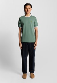 Giovane uomo in piedi, che indossa una t-shirt verde a maniche corte con bordo bianco, pantaloni neri e scarpe in suede beige, su uno sfondo neutro.