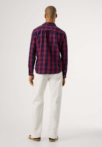 Camisa xadrez em azul-marinho e vermelho, com mangas compridas e punhos com botão. Calças brancas de corte reto, calçado casual, fundo plain.