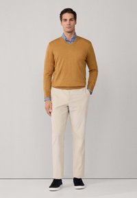 Uomo che indossa un maglione a V color senape sopra una camicia a righe blu, pantaloni beigi e sneaker nere, in piedi con una mano in tasca contro un muro grigio.