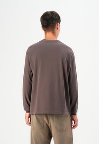 Cămașă cu mânecă lungă, de culoare maro, confecționată dintr-un material moale, cu guler rotund și o croială lejeră, asortată cu pantaloni de culoare maro deschis.