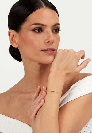 Guld kædearmbånd med røde, lilla og klare perler, båret på håndleddet mod en neutral baggrund; glat tekstur og delikat design.