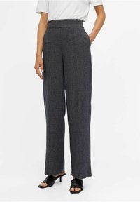 Pantalons gris taille haute à jambes larges avec une texture subtile. Présente des poches latérales et un design épuré, associés à des escarpins noirs à bout ouvert.