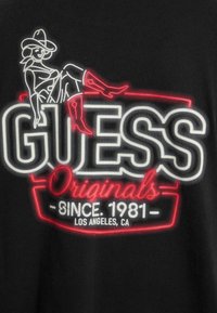 T-shirt nero con un design grafico al neon che presenta la parola "GUESS", un disegno a linee giocoso di una figura e dettagli testuali in rosso e bianco.