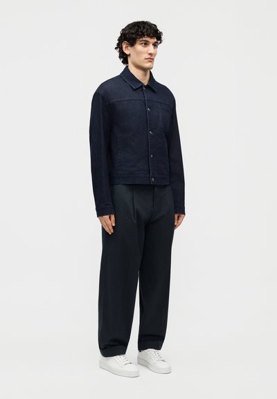 Göndör sötét hajú férfi sötét farmerdzsekiben, bő fekete nadrágban és fehér tornacipőben, egyszerű háttér előtt állva.