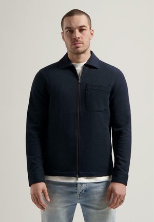 Giacca a zip blu navy in materiale testurizzato; presenta un colletto e una tasca sul petto. Indossata sopra una camicia bianca e abbinata a jeans azzurri chiari.