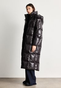 Caban lung, lucios, din material pufos, cu glugă, având un design matlasat și închideri cu capse. Asortat cu blugi negri cu picioare largi și încălțăminte neagră.