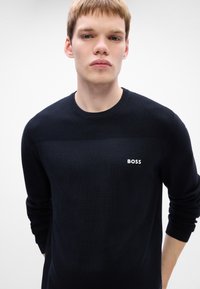 Tânăr cu părul scurt și blond, îmbrăcat într-un pulover negru cu logo-ul "BOSS", stând în fața unui fundal alb simplu.