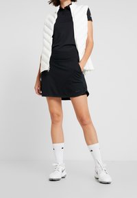 White puffer vest, black polo, black pleated skirt, white socks with black accents, and white Nike shoes, set against a plain background.