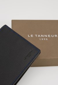 Black leather wallet with blue stitching, displaying a textured surface, resting on a brown packaging box with "LE TANNEUR 1898" printed on it.