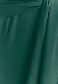 Tamsiai žalia medžiaga su lygiu paviršiumi, turinti krentanti raukšles ir lengvą blizgesį. Paprastas dizainas be matomų raštų ar aparatūros.