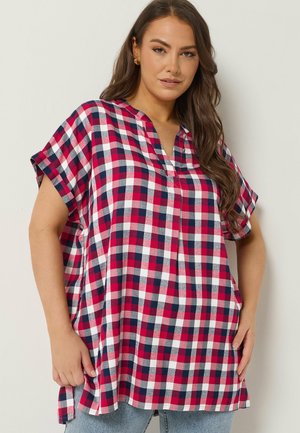 Frau trägt ein locker sitzendes kurzärmeliges Oberteil mit V-Ausschnitt in rot, weiß und blau kariert, steht vor einem schlichten hellen Hintergrund.