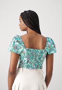 Zelený kvetinový crop top s nariasenými rukávmi, nariaseným pásom a štvorcovým výstrihom. Textúra je hladká; vzor obsahuje ružové a biele akcenty.